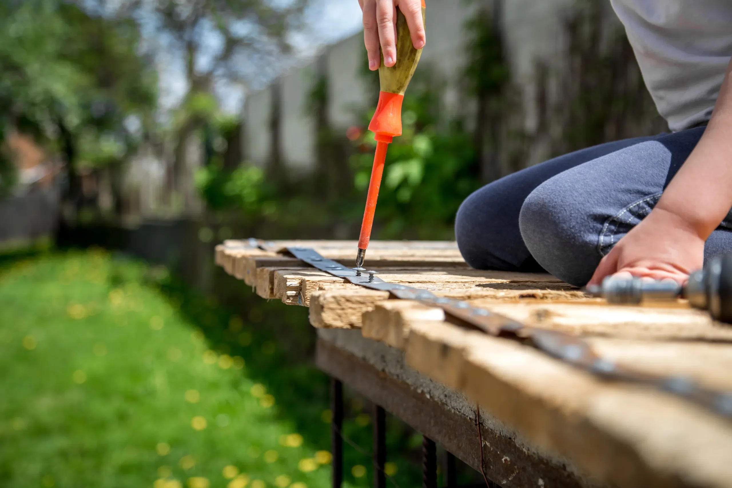 Child using a cordless power drill to fasten screws into a wooden pallet outdoors.