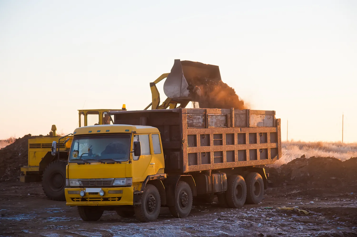 Tracked excavator loading soil into a large dump truck on a site during early morning sunrise.
