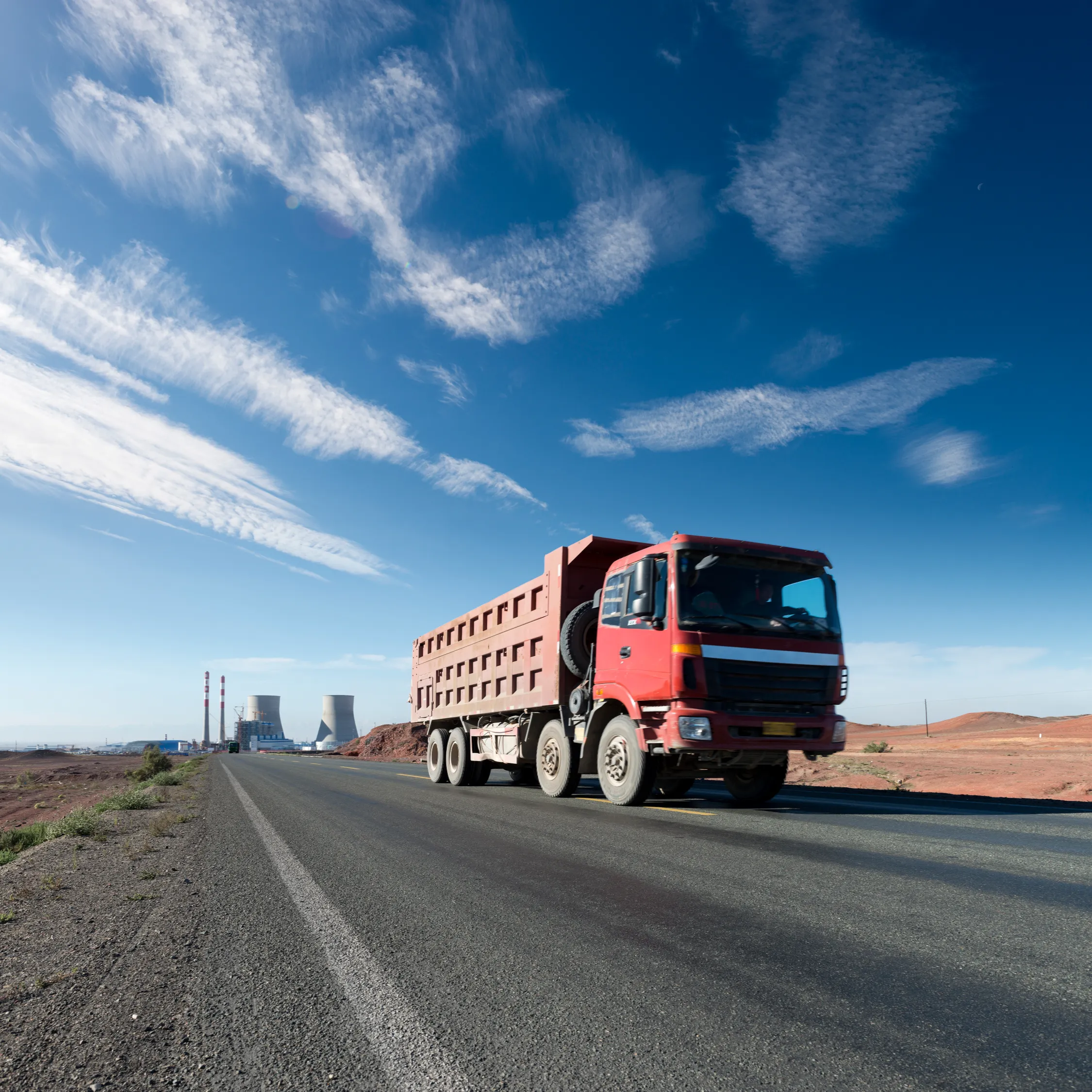 Red semi-truck hauling a heavy flatbed trailer along an open asphalt highway under a clear blue sky.